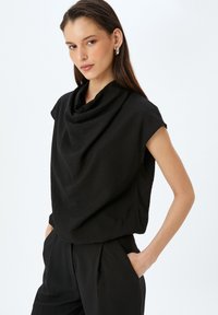 Sort draperet top med cowl-udskæring og ærmer med cap-sleeves. Blødt, tekstureret stof. Vist sammen med matchende sorte bukser.