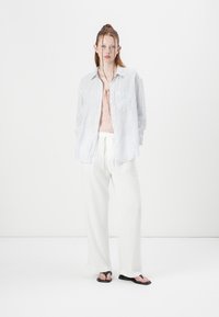 Gestreepte blauwe en witte button-up shirt, lichtroze camisole, witte wijde broek en zwarte flip-flops, geplaatst tegen een effen achtergrond.