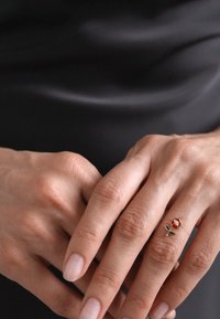 Anello d'argento con un piccolo design di rosa rossa e un gambo verde, indossato su una mano con unghie naturali su uno sfondo scuro.