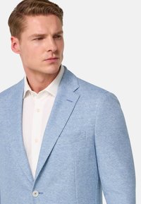 Blazer azul claro con tejido texturizado, solapas de muesca, cierre de un botón y un bolsillo en el pecho, llevado sobre una camisa blanca.
