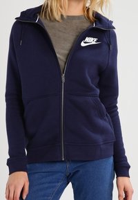 Námorně modrá zipová mikina s kapucí a bílým logem Nike na levé hrudi, má kapsy a žebrované manžety, vyrobena z měkké, strukturované tkaniny.