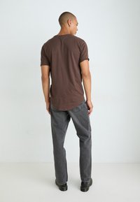 Brown short-sleeve t-shirt with a round neckline, paired with gray straight-leg jeans and black lace-up boots, seen from the back.