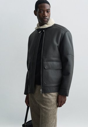 Schwarze Lederjacke mit Schaffellkragen, Frontreißverschluss und zwei großen Taschen. Trägt man über einem dunklen Pullover und strukturierten hellbraunen Hose.