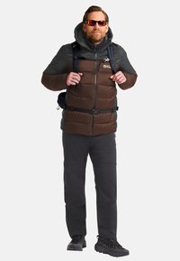 Uomo che indossa occhiali da sole, giacca a vento marrone e grigia, pantaloni da escursionismo neri, scarponi e che porta uno zaino con fascia toracica.