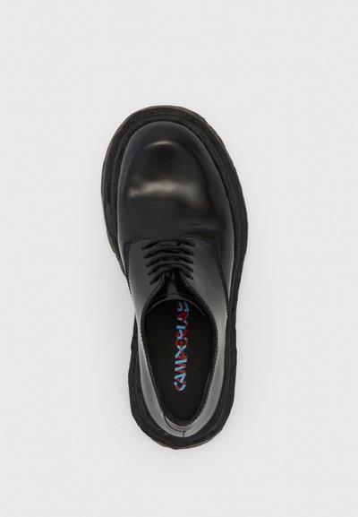 Black leather shoe with a smooth texture, featuring a rounded toe and central lacing. Thick rubber sole provides a contrasting, cushioned edge.