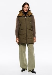 Chaleco acolchado largo en color verde olivo con capucha desmontable, que cuenta con bolsillos frontales con cremallera y un exterior suave. Combinado con pantalones de pierna ancha en un tono verde claro.