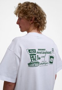 Balta medvilninė marškinė su žalia grafika ant nugaros. Dizainas apima restoranų ženklus ir įvairius teksto elementus. Atsipalaidavęs siluetas.