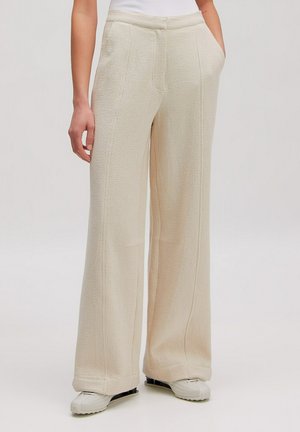 Person steht und trägt beige Hose mit hohem Bund und weitem Bein sowie seitlichen Taschen und weiße Sneakers vor einfarbigem Hintergrund.