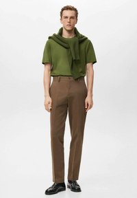 Muž v olivovo zelenej krátkosúčkej košeli s rovnakým svetrom prehodeným cez ramená, hnedé nohavice a čierne topánky stojaci na bielom pozadí.