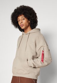 Felpa beige con cappuccio realizzata in cotone, con tasca frontale, polsini a coste e una cerniera rossa con dettaglio del cartellino sulla manica.