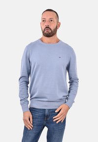 Uomo con barba che indossa un maglione a maniche lunghe blu chiaro e jeans blu, in piedi con una mano vicino alla tasca contro uno sfondo bianco.