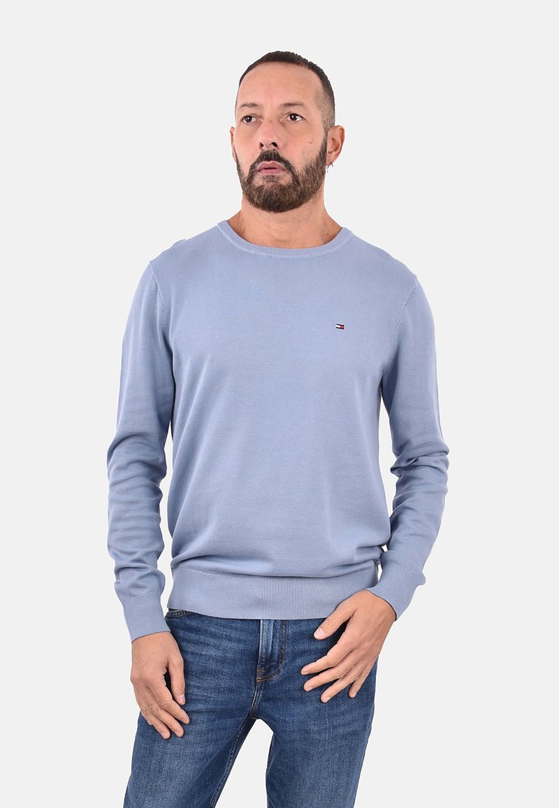 Uomo con barba che indossa un maglione a maniche lunghe blu chiaro e jeans blu, in piedi con una mano vicino alla tasca contro uno sfondo bianco.