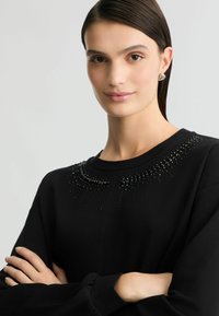 Zwarte sweater met een ronde hals, voorzien van een kraalversiering langs de kraag. Zachte textuur en een ontspannen pasvorm.