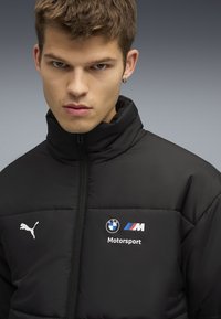 Sort pufferjakke med høj krave, med hvidt Puma-logo og BMW Motorsport-branding på brystet. Glat tekstur, lynlåslukning.