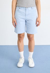 Shorts en denim bleu clair avec une coupe droite, cinq poches et fermeture à bouton, associés à des baskets blanches et des chaussettes à mi-mollet.