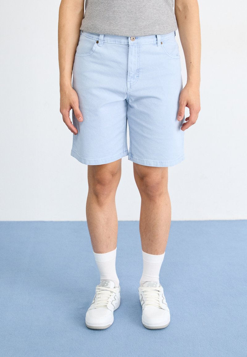 Shorts en denim bleu clair avec une coupe droite, cinq poches et fermeture à bouton, associés à des baskets blanches et des chaussettes à mi-mollet.