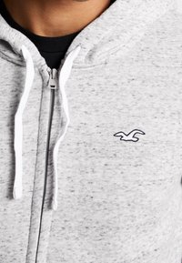 Felpa con zip grigia con texture screziata, cordini bianchi e logo di un uccello ricamato in blu navy sul petto sinistro.