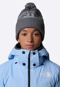 Giacca impermeabile azzurro chiaro con bottoni e zip neri, abbinata a un berretto in maglia grigia con pon-pon e logo a contrasto.