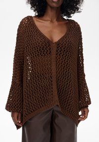 Brązowy sweter dzianinowy z dekoltem w serek, otwartym splotem, długimi rękawami i asymetrycznym dołem, w zestawieniu z skórzanymi spodniami.