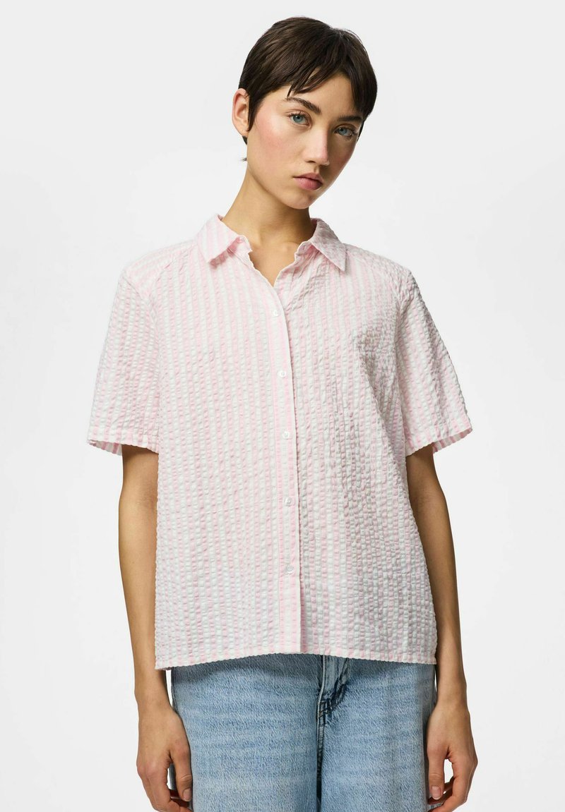 Jeune personne aux cheveux courts et foncés portant une chemise légère rose pâle à manches courtes texturée et un jean bleu clair.
