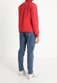 Personne portant une veste matelassée rouge, un jean bleu et des baskets montantes blanches avec un motif à carreaux noir, se tenant de dos.