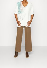 Top blanco de cuello en V con diseño abstracto en tonos verde azulado y rosa; combinado con pantalones de pierna ancha marrones y botas de tobillo marrones. Textura suave en general.