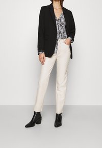 Sort blazer med revers, kombineret med en sort og hvid mønstret bluse, cremefarvede bukser og sorte ankelstøvler. Ensfarvet baggrund.