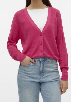 Mujer con un cárdigan de botones color rosa brillante sobre una camiseta blanca y jeans de talle alto en azul claro, con la mano en el bolsillo.