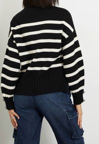 Maglione a strisce bianche e nere con orlo e polsini a coste, dotato di bottoni sulle maniche, indossato sopra pantaloni cargo blu.