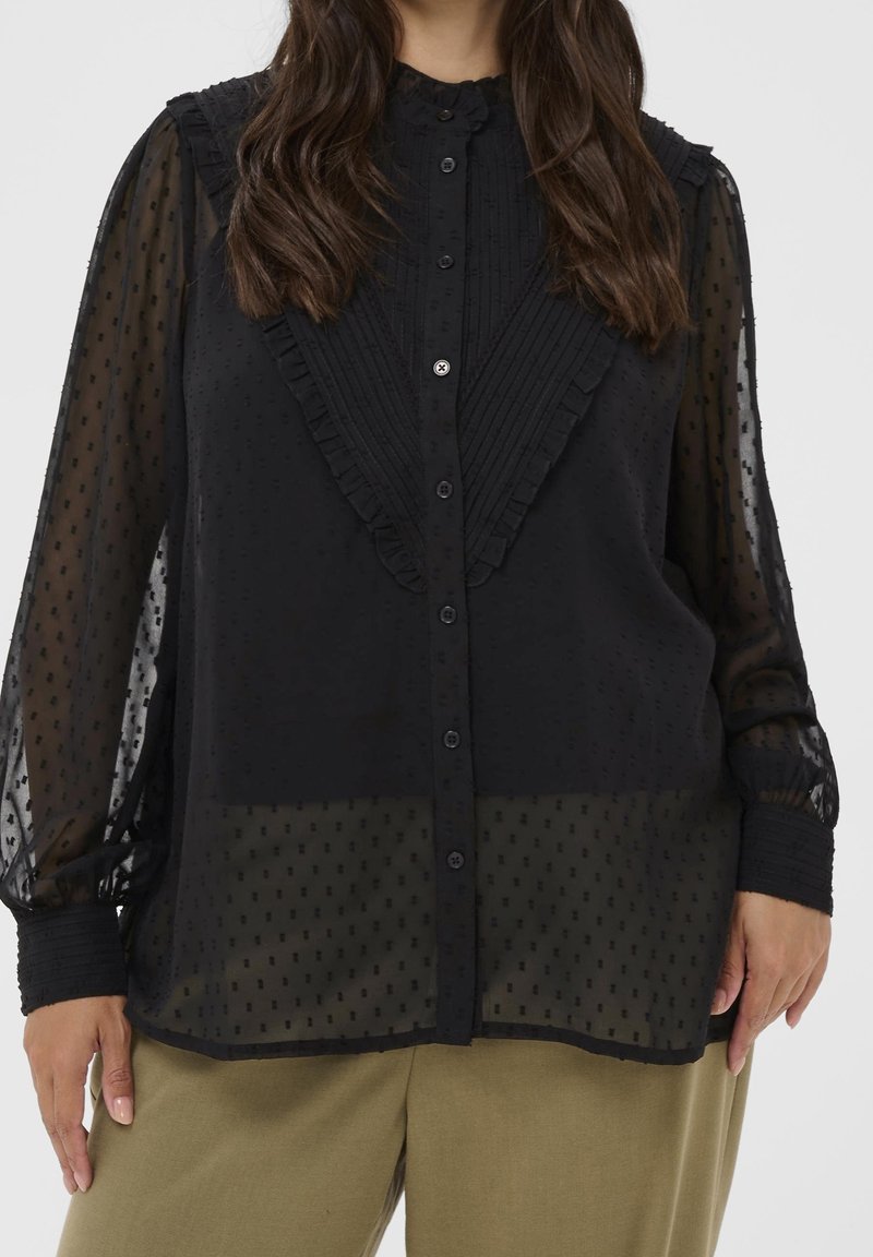 Femme portant un chemisier noir transparent à manches longues avec des détails à volants, associé à un pantalon marron clair, sur un fond uni.
