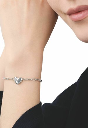 Braccialetto con ciondolo a forma di cuore in argento indossato al polso, con il braccio appoggiato vicino al volto, persona vestita di nero con carnagione chiara.