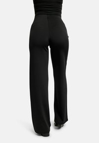 Žena v čiernych širokých nohaviciach s vysokým pásom a čiernom topánke stojí chrbtom k svetlému jednoliatemu pozadiu.