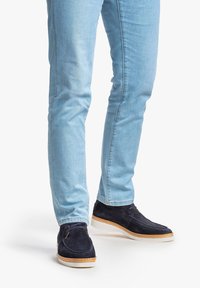 Scarpe scure in suede con suola beige, abbinate a jeans slim-fit azzurri. Le scarpe presentano cuciture minime e lacci.