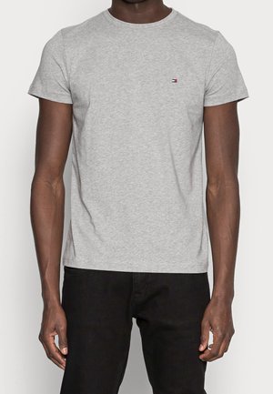 Mann trägt ein schlichtes hellgraues kurzärmeliges T-Shirt mit kleinem Logo auf der Brust und schwarze Hose, steht mit entspannten Armen an den Seiten.