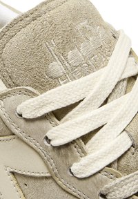 Sneaker beige in suede con lacci intrecciati bianchi, dettaglio del logo in rilievo e superficie superiore testurizzata con cuciture in contrasto.