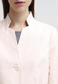 Blazer rosa claro con cuello mao, tejido texturizado y cierre de un solo botón en el frente. Parte de una camiseta gris es visible por debajo.