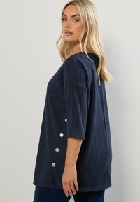 Maglione a coste navy con maniche a tre quarti e dettagli di bottoni sui lati. Tessuto morbido con una vestibilità comoda e un orlo arrotondato.