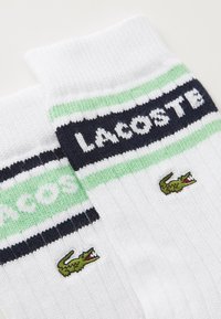 Witte geribde sokken met groene en marineblauwe strepen, met "LACOSTE" geborduurd op de boord en een klein groen krokodillenlogo.