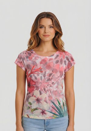 Giovane donna con indosso una t-shirt rosa a maniche corte con motivi floreali e foglie, in piedi davanti a uno sfondo semplice.