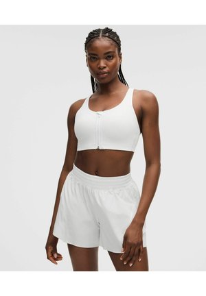 Frau mit geflochtenem Haar, die einen weißen Sport-BH mit Reißverschluss vorne und weiße Sportshorts trägt, steht vor einem einfarbigen Hintergrund.
