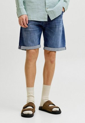 Personne portant un short en jean bleu avec revers, des chaussettes crème, des sandales marron et une chemise à manches longues vert clair, main dans la poche, debout sur fond blanc.