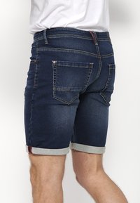 Homme portant un short en denim bleu foncé retroussé et une chemise blanche, vu de dos sur un fond blanc uni.