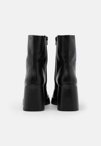 Une paire de bottines en cuir noir avec des talons blocs et des fermetures éclair à l'arrière, vues de l'arrière sur un fond blanc.