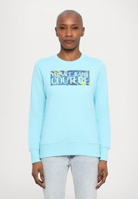 Világoskék pulóver, amelyen egy téglalap alakú grafika található, rajta a "Versace Jeans Couture" felirattal fehér és sárga színben, egy mintás háttéren.