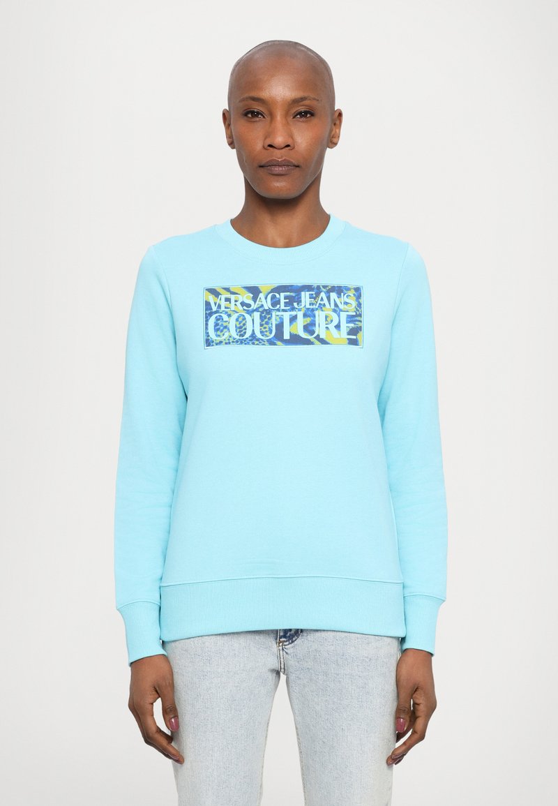 Világoskék pulóver, amelyen egy téglalap alakú grafika található, rajta a "Versace Jeans Couture" felirattal fehér és sárga színben, egy mintás háttéren.
