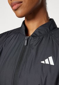 Črna zip-up jakna iz lahkega materiala, ki ima visok ovratnik in bel trikotni logotip na desni strani.