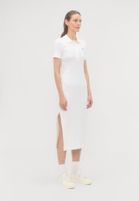 Biele polo šaty s krátkymi rukávmi, gombíkovou légou, bočnými rozparkami a logom Lacoste. Spárované s bielymi teniskami a členkovými ponožkami.