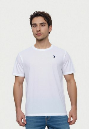 Ung mand iført hvid kortærmet T-shirt med lille broderet logo og blå jeans, stående mod en ensfarvet lys baggrund.