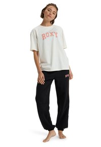 Bílá oversized tričko s logem "ROXY" v korálové barvě. Černé tepláky s logem "ROXY". Uvolněný střih. Pohodlný materiál z bavlněné směsi.