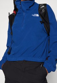 Niebieski polarowy sweter z wysokim kołnierzem i zapięciem na pół zamka. Posiada elastyczny dół i mankiety; logo na klatce piersiowej. Widoczne czarne paski plecaka.